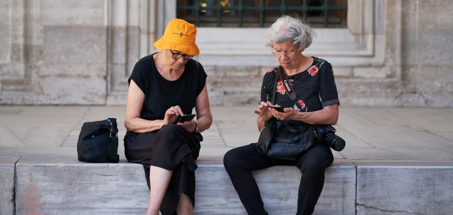 Zwei Seniorinnen sitzen auf einer Mauer und haben Smartphones in der Hand. Eine Frau hat einen Fotoapparat umhängen.